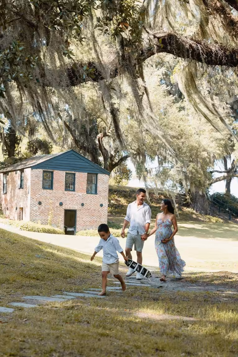 Parents and a young boy in white and a long floral dress walking on a path under moss-draped live oaks near a small brick building