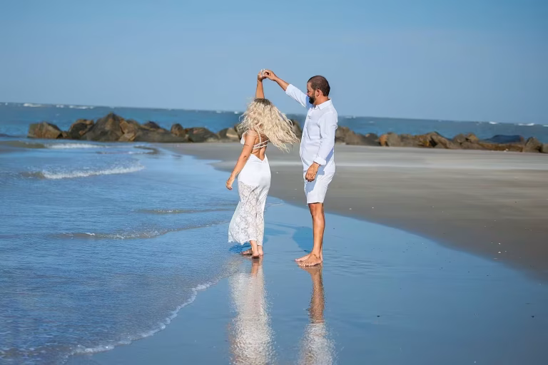 Couple in white on a beach at the water's edge, the woman spinning under the man's raised hand, their reflections on the wet sand and a rock jetty behind them