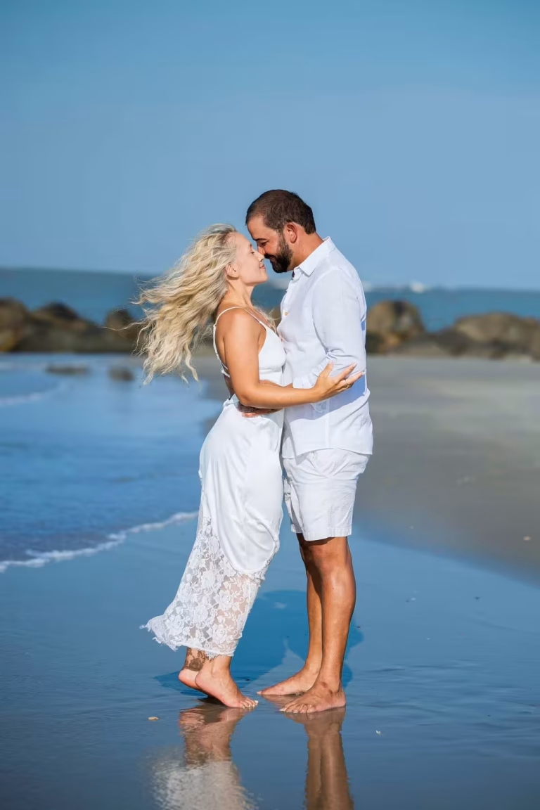 Couple in white clothes embracing nose to nose at the water's edge on a sunny beach, with rocks along the shore behind them