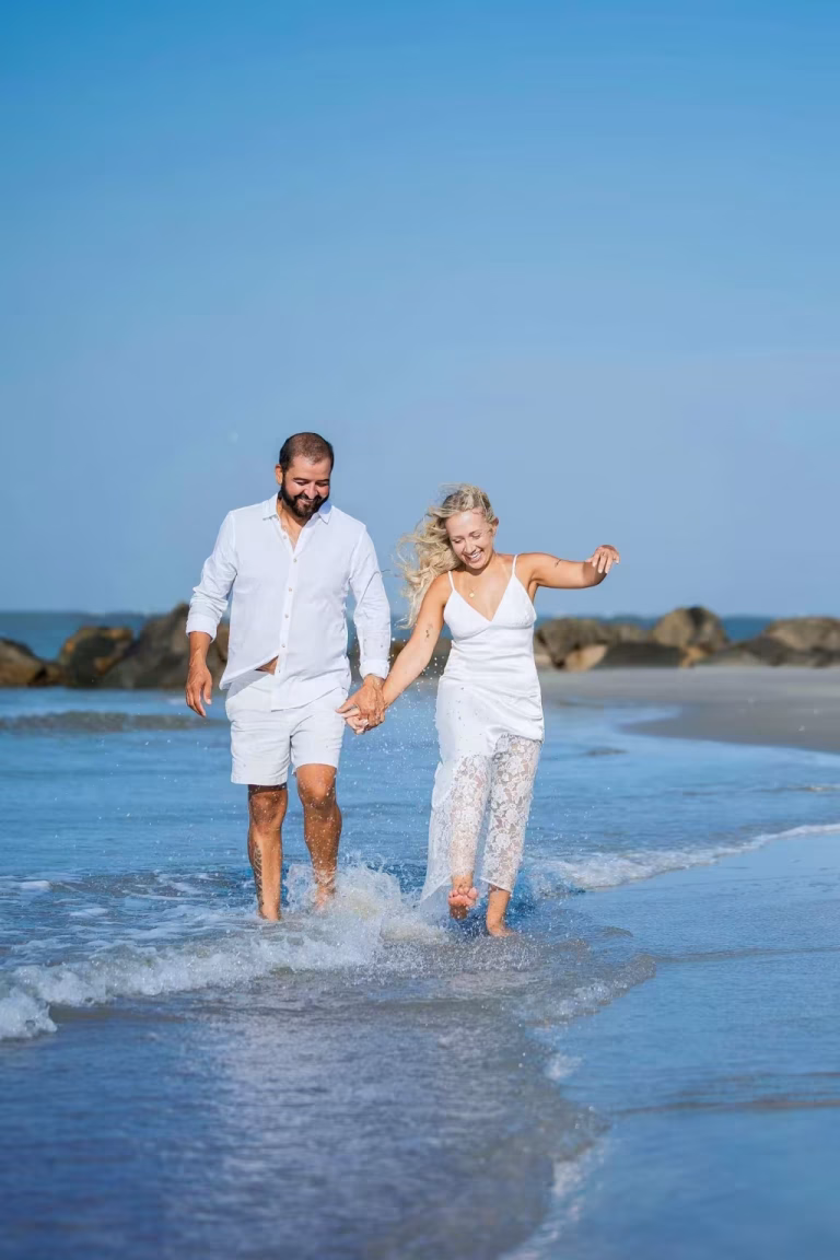 Couple in white linen and lace walking hand in hand through ankle-deep surf, laughing, with a rock jetty and blue sky behind them