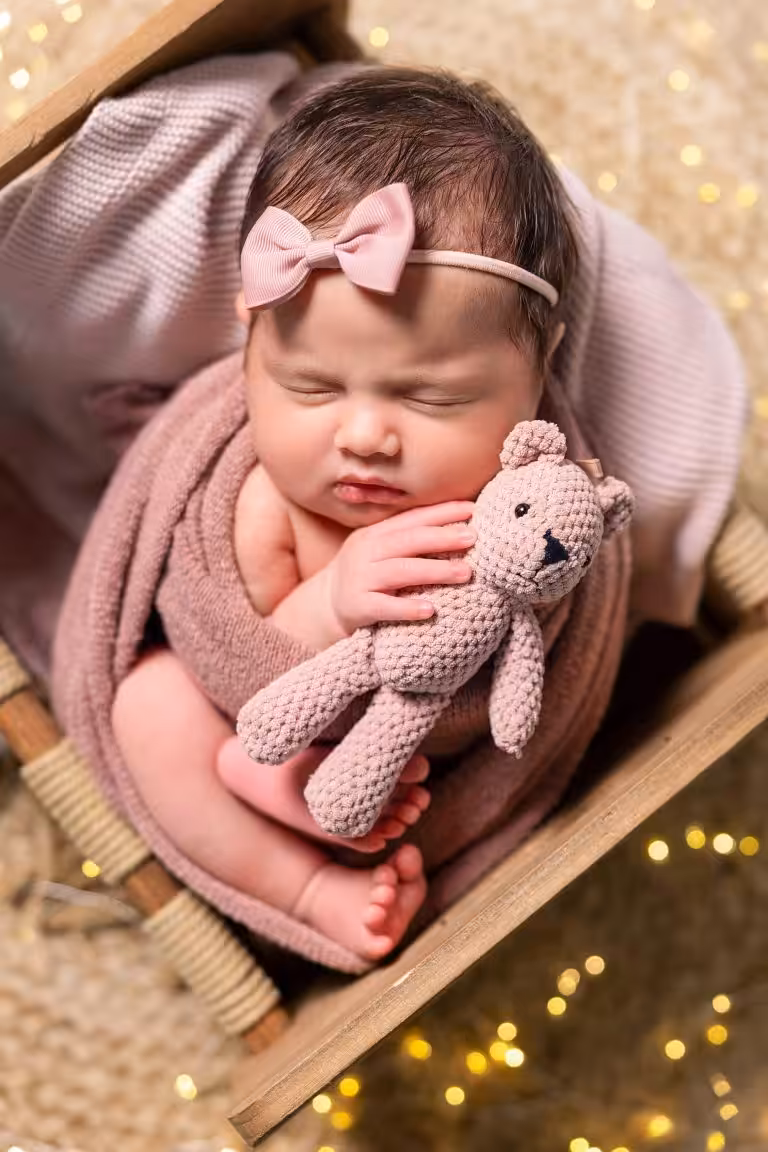 Christmas Family Photo Ideas for a December Without Snow Sleeping newborn wrapped in a pink knit blanket with a bow headband and a knitted teddy bear, in a small wooden crate with soft golden lights blurred behind