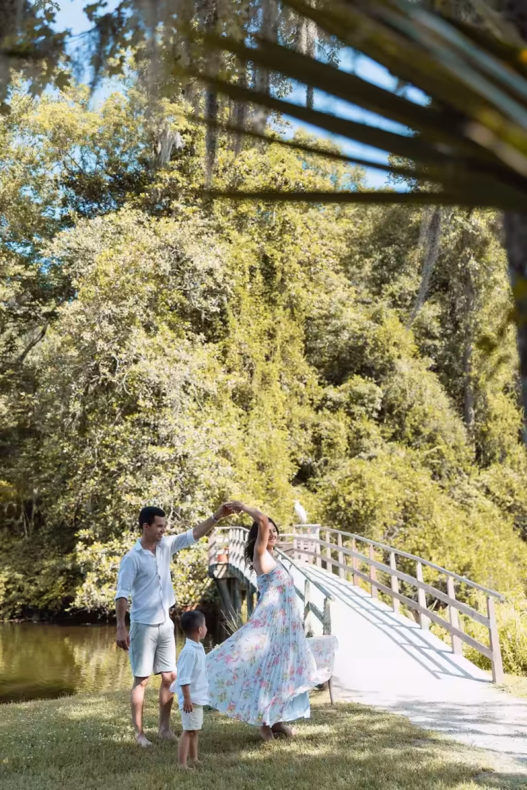 Father, young son and mother in a flowing floral dress dancing on a lawn beside a pond and a white wooden footbridge under trees with Spanish moss