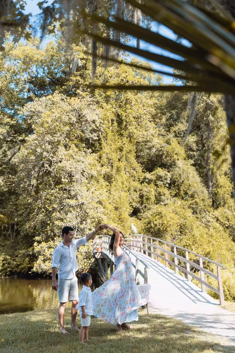 Family Photo Poses: A Reference Guide From Real Sessions Father in a light shirt twirling the mother in a long blue floral dress beside a white wooden footbridge over a pond, while their small son watches
