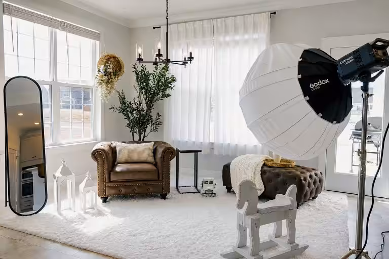 Studio set with a brown leather armchair, tufted ottoman, white rocking horse, tall mirror and sheer-curtained windows, with studio lights standing in the room