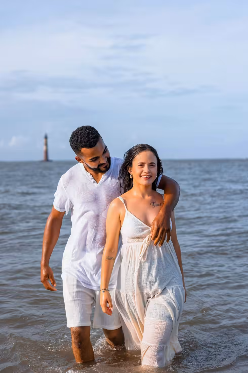 Engagement Photo Ideas, Chosen by Where the Photos Will End Up Couple in white clothes wading knee-deep in calm water, his arm around her shoulders, with a lighthouse standing offshore on the horizon