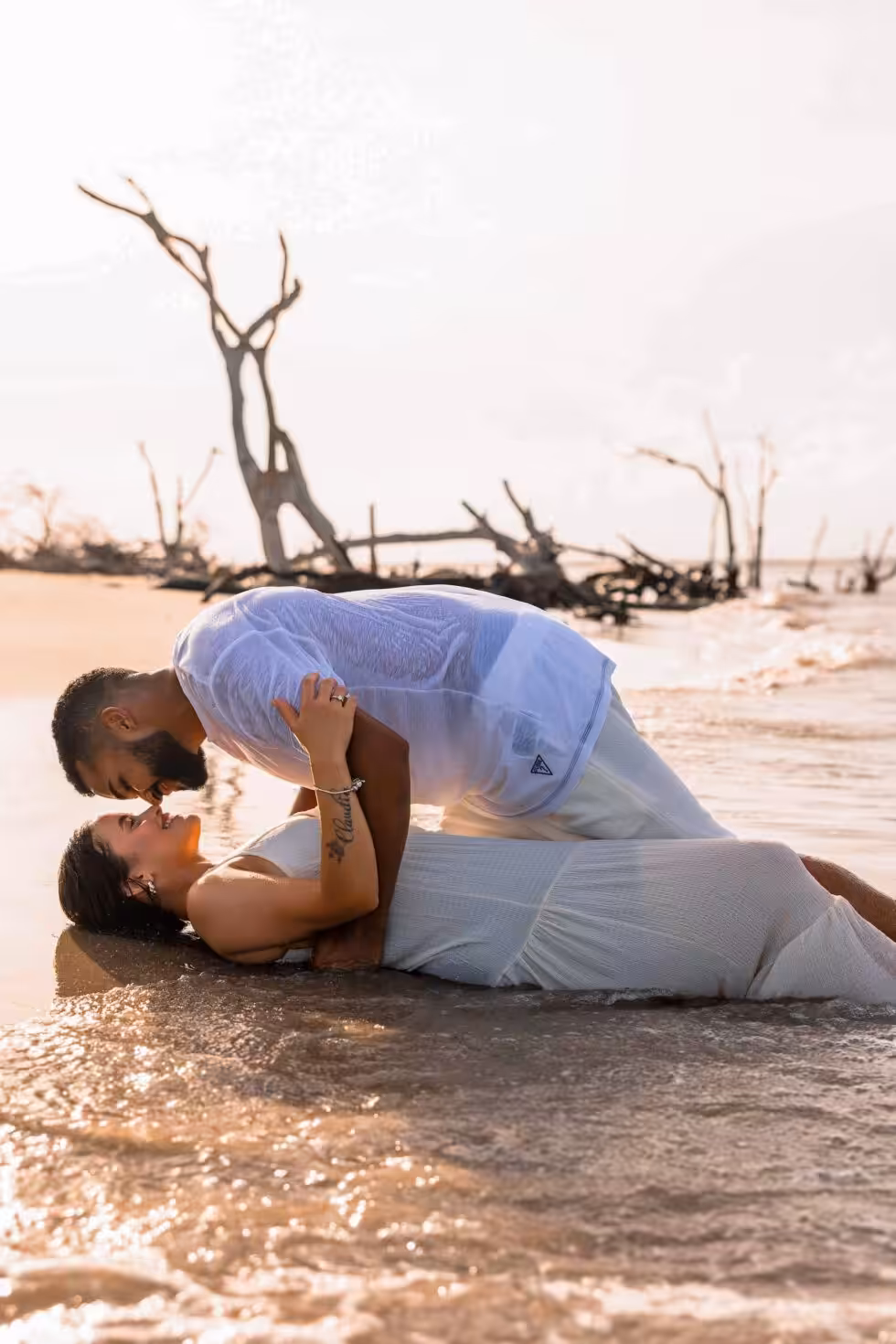 Engagement Photo Ideas, Chosen by Where the Photos Will End Up Couple lying at the waterline on a beach, a wave washing around them, with bleached driftwood trees behind