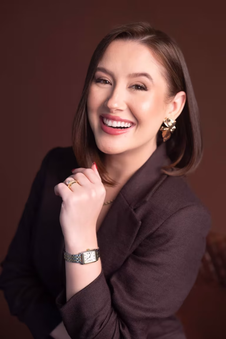Woman in a dark brown blazer laughing with one hand raised near her collar, against a brown studio backdrop