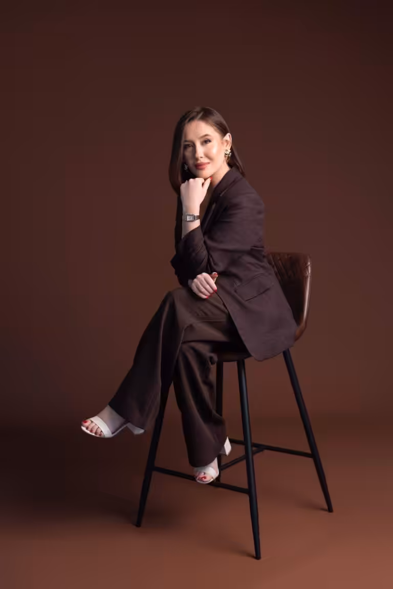 Woman in a dark brown suit sitting on a tall black stool against a deep brown studio backdrop