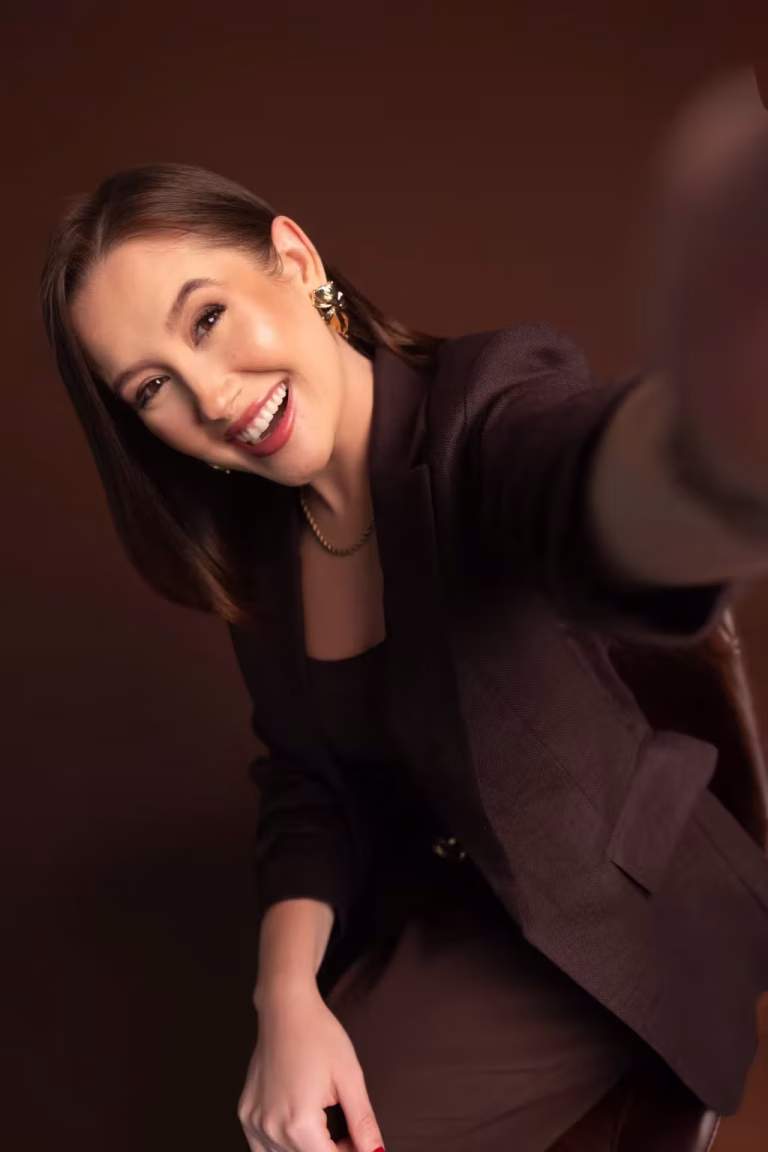 Woman in a dark brown suit laughing and leaning toward the camera, one hand reaching toward the lens out of focus, against a chocolate-brown backdrop