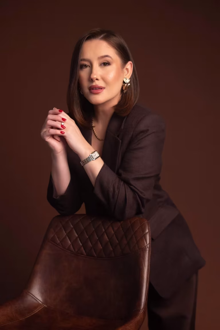 Woman in a dark brown blazer leaning on the back of a leather chair with her hands clasped, looking at the camera with a small smile, against a plain brown studio backdrop