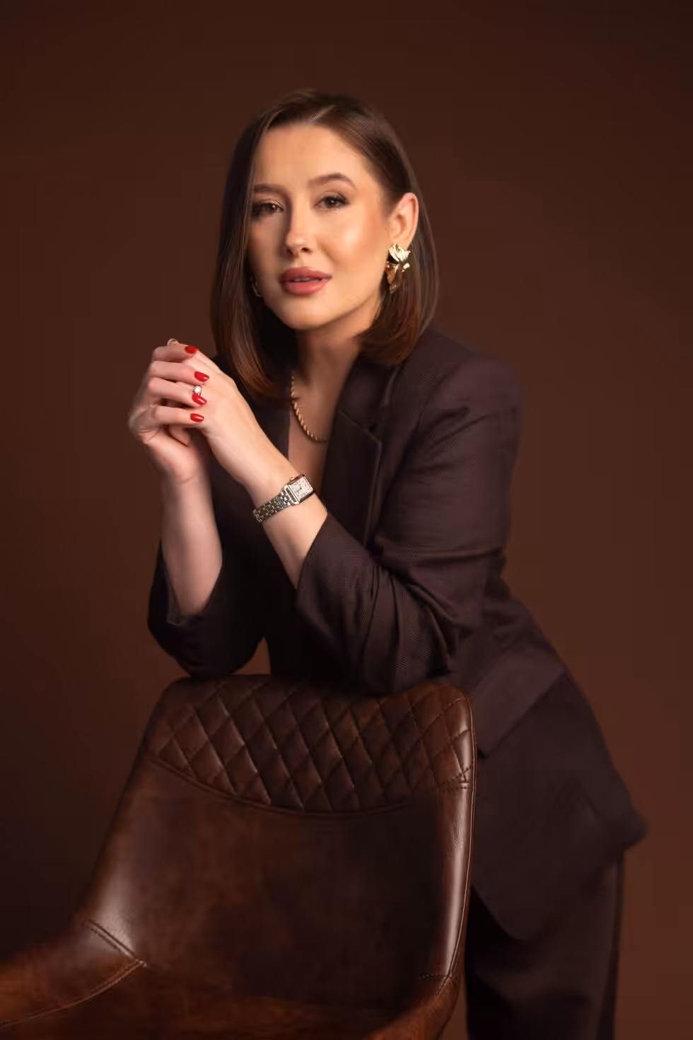 How to Prepare for a Headshot: Decide These Five Things First Woman in a dark brown blazer and trousers leaning on the back of a leather chair with her hands clasped, against a brown studio backdrop