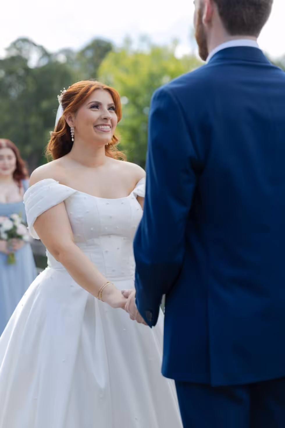 First Look Wedding Photos: Should You See Each Other Before the Ceremony? Bride in an off-shoulder gown smiling at the groom, seen from behind in a navy suit, as they hold hands during the ceremony, with a bridesmaid in blue behind her