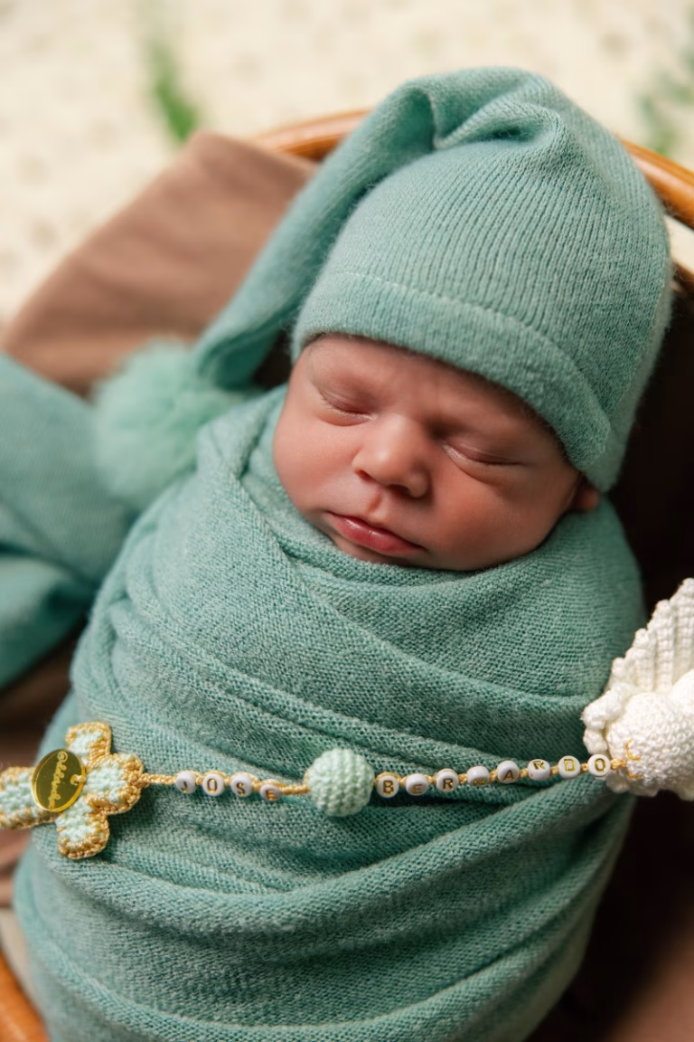 Newborn boy asleep in a round woven basket, swaddled in a green knit wrap with a matching sleepy cap and a beaded keepsake across the wrap