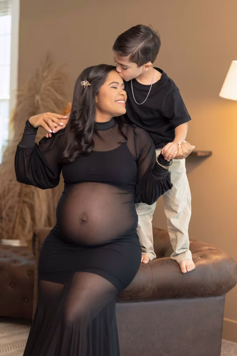 Pregnant woman in a sheer black gown sitting on the edge of a leather sofa while her young son, standing on the seat, kisses the top of her head