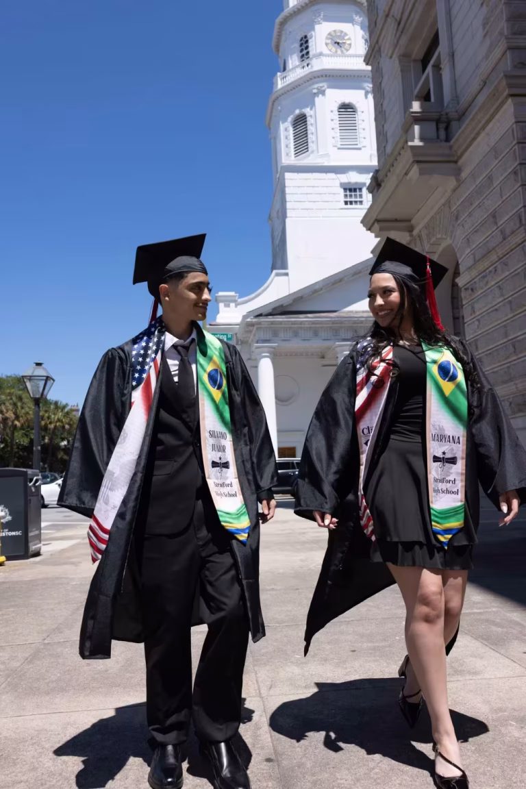 Two graduates in open black gowns and stoles walking down a downtown Charleston street, a white church steeple behind them