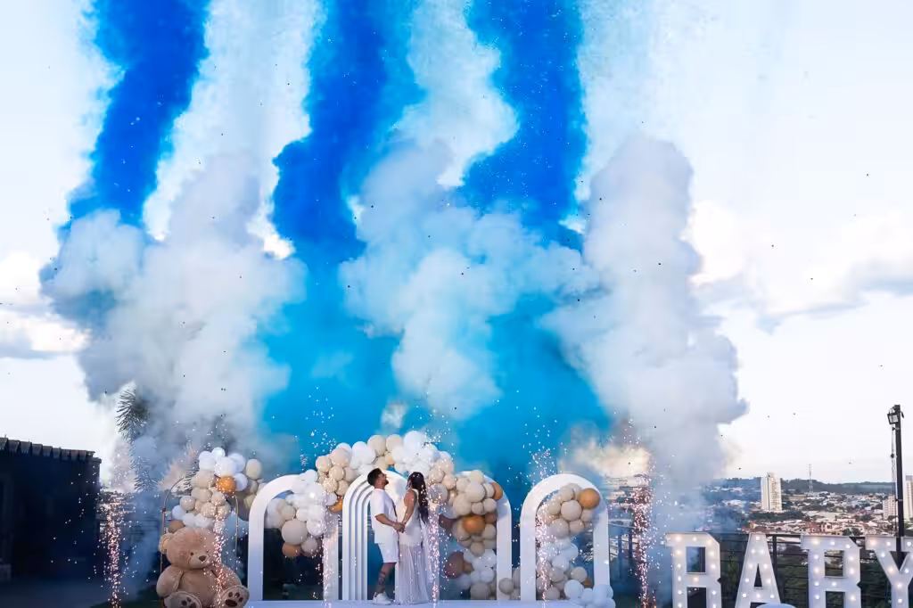 Gender Reveal Ideas, Chosen by Who Is There and Where It Happens Columns of blue smoke rising behind a stage of white arches, balloons and a large teddy bear, with the expecting couple standing in the center