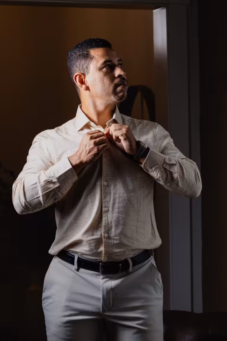 How to Prepare for a Photoshoot, Step by Step Man in a beige button-up shirt and gray trousers fastening his cuff, lit from the side against a dark background