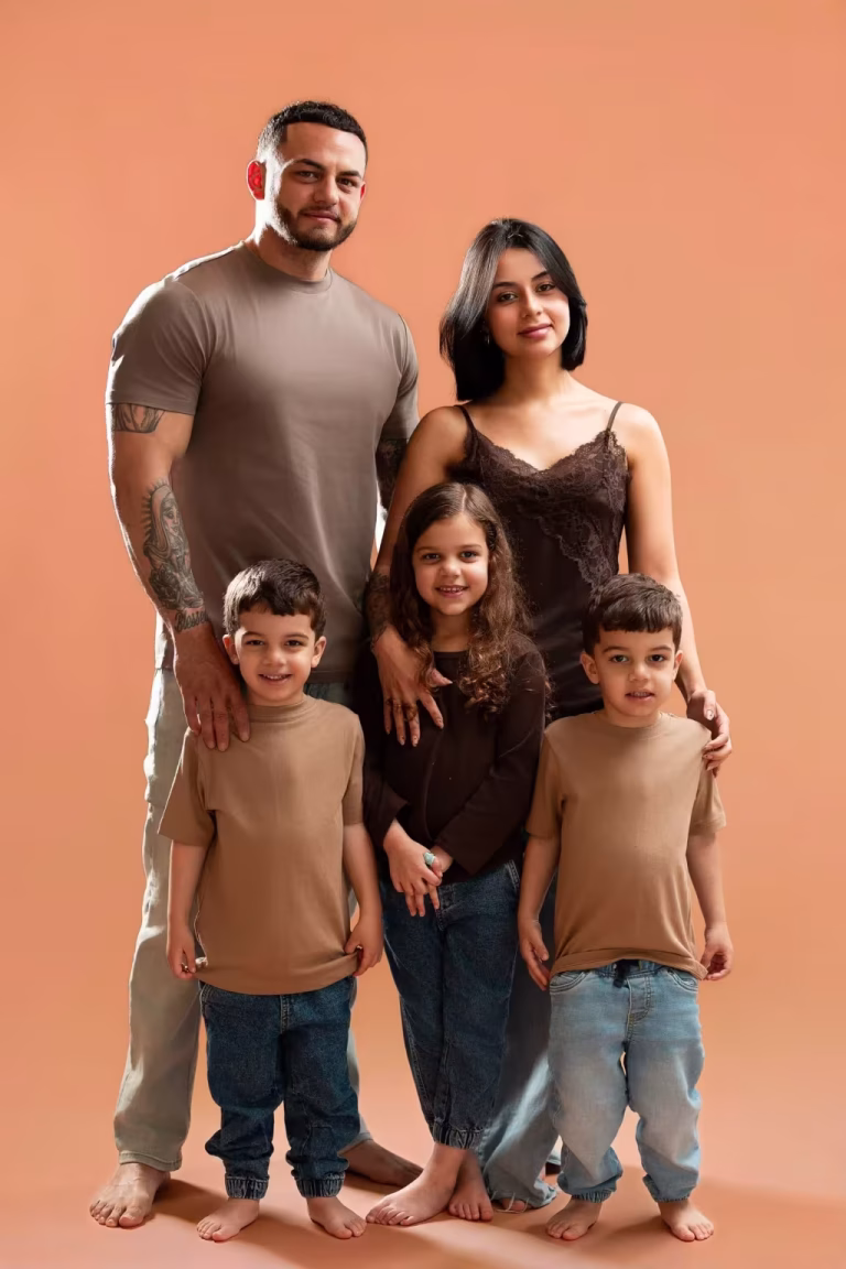 Family of five standing barefoot against a peach studio backdrop, dressed in brown, taupe and black tops with blue jeans