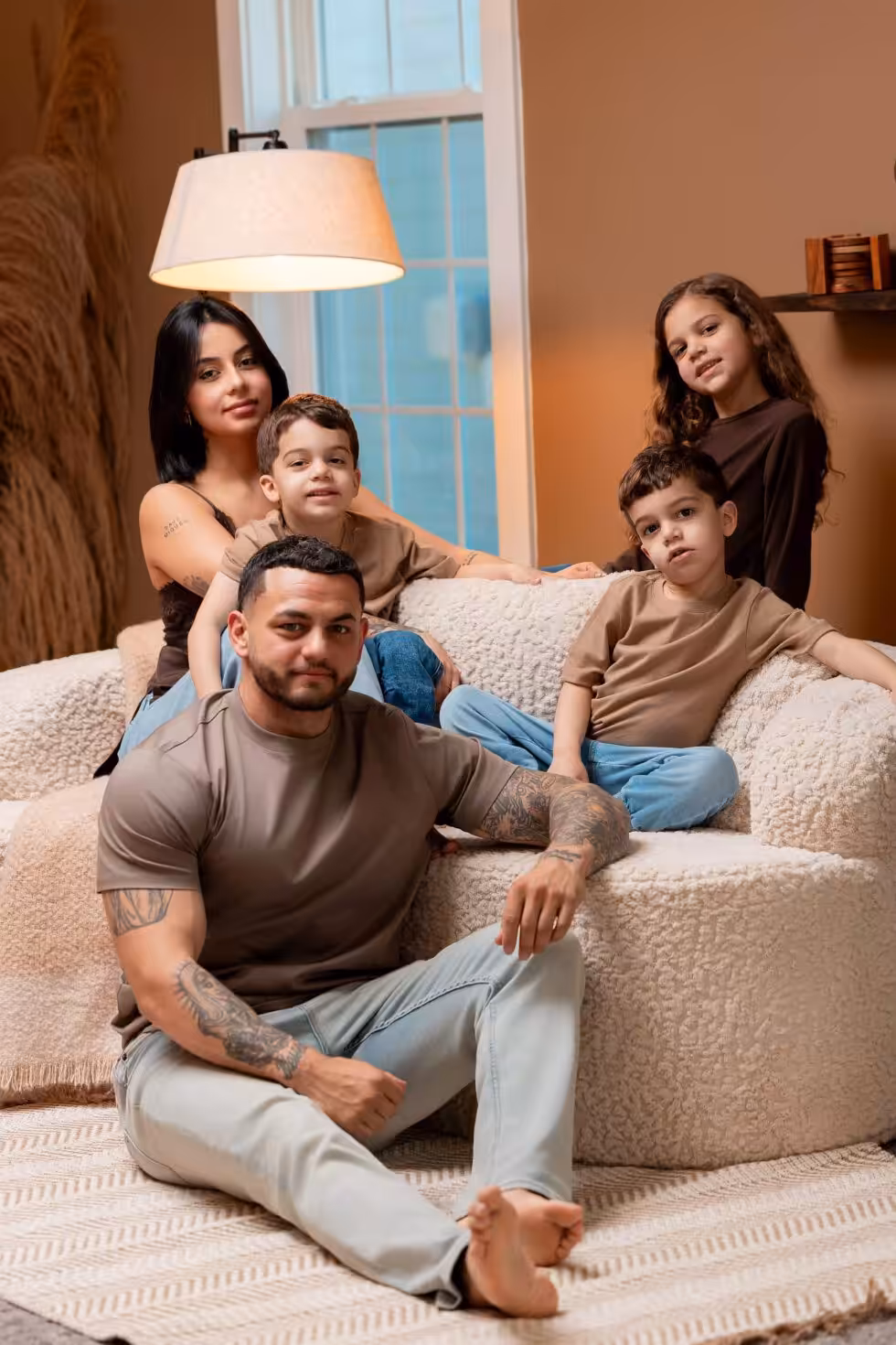 Family Photo Poses: A Reference Guide From Real Sessions Father sitting on a rug in front of a cream boucle sofa while the mother and three children sit and kneel on the sofa behind him, under a warm floor lamp