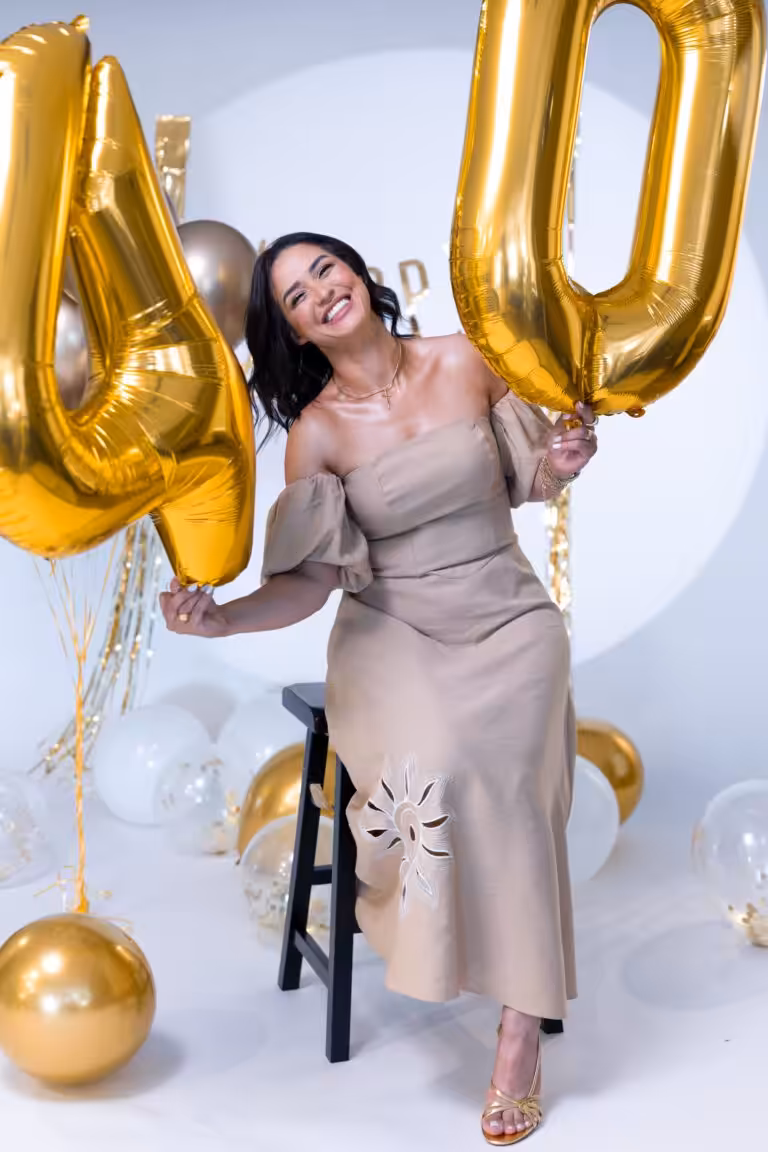 Birthday Photoshoot Ideas: How a Real 40th Came Together, Set by Set Woman in a beige off-shoulder dress holding giant gold number 4 and 0 balloons in a white studio set with gold streamers