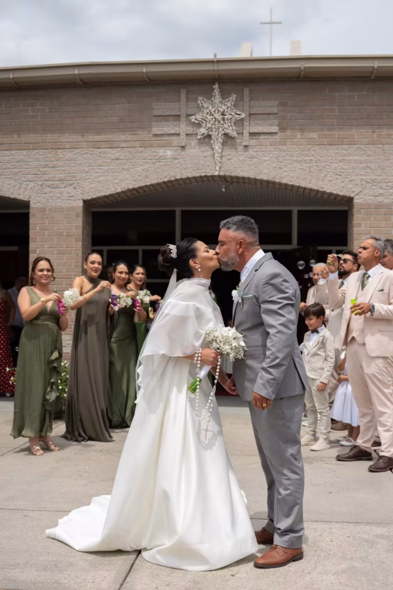 Bride and groom kissing outside the church doors as guests and bridesmaids in sage green dresses cheer around them