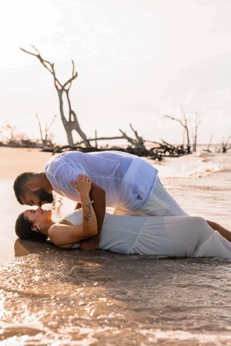 Couple in white lying at the water's edge on a beach, with weathered driftwood trees behind them