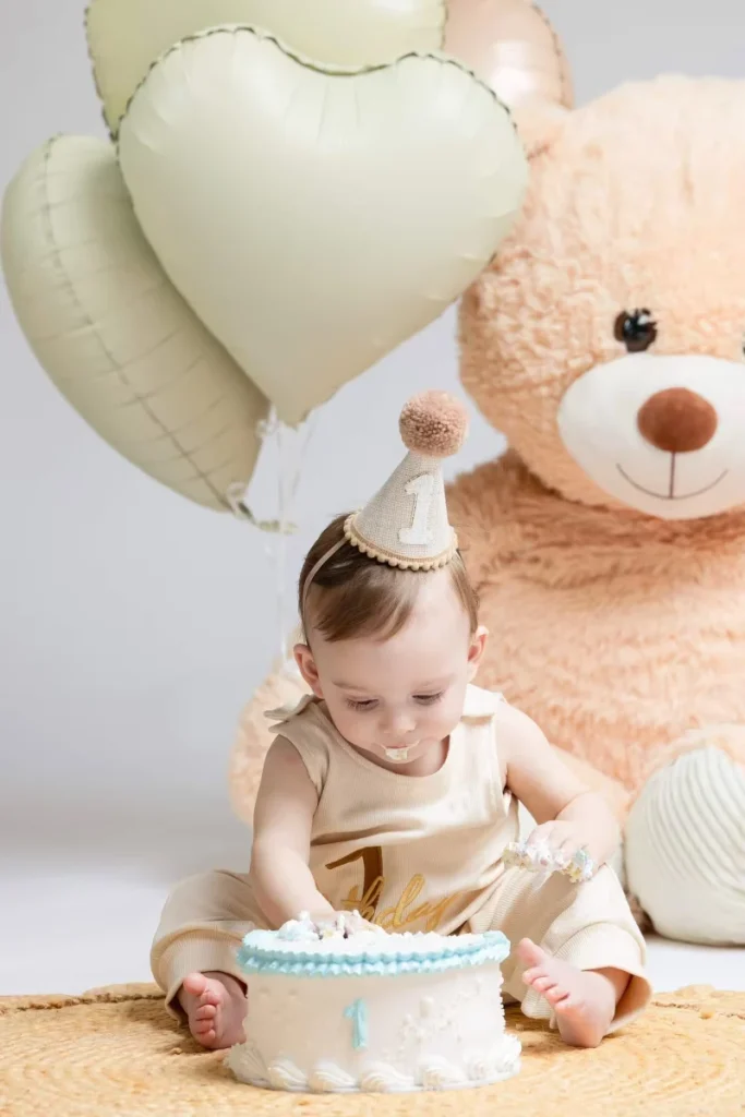 Baby in a party hat sitting on a woven rug with a small cake, a large teddy bear and cream balloons in a studio set
